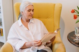 Older senior woman reading book, about flooring options for seniors, in a yellow chair.